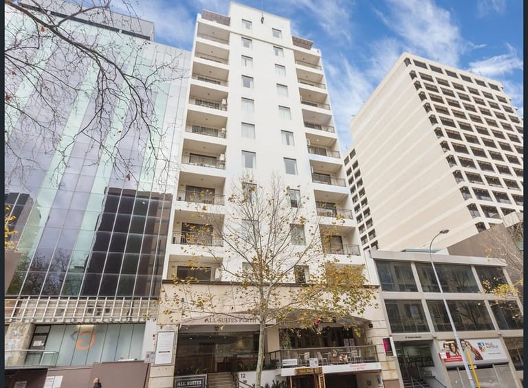 Urban street view showing multi-story residential and commercial buildings with modern white facades and large windows under a blue sky.